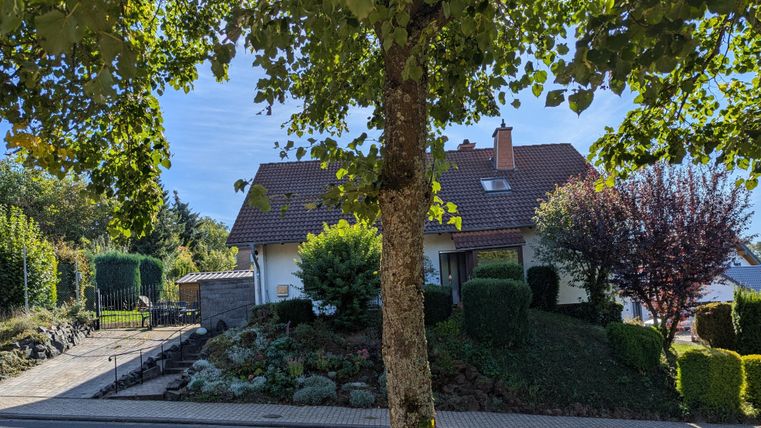 Een klein huis omringd door bomen en een onderhouden tuin. De lucht is helder en zonnig.