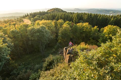 Zwei Personen stehen auf einem Felsen mit Blick auf einen Wald und Hügel im Hintergrund.