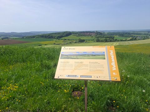 An information sign stands on a green meadow overlooking the hilly landscape. In the background, forests and fields can be seen.