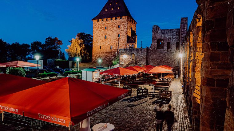 Ein gemütliches Restaurant im Freien mit roten Sonnenschirmen und einer historischen Burg im Hintergrund. Die Szene ist bei Nacht beleuchtet und vermittelt eine einladende Atmosphäre.