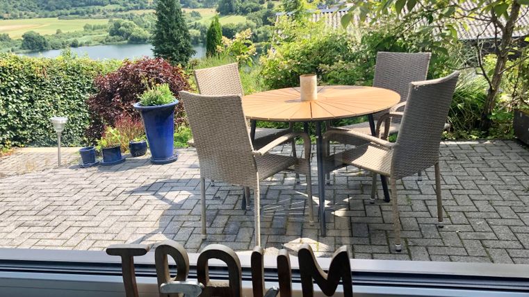 Een gezellige buitenruimte met een ronde tafel en stoelen. Op de achtergrond strekt zich een groene landschap uit met zachte heuvels en een rivier.