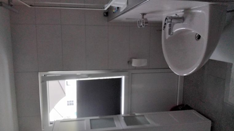 Ein modernes Badezimmer mit einem Waschbecken, einer Dusche und einem Fenster. Die Wände sind hell gefliest und der Boden ist grau.