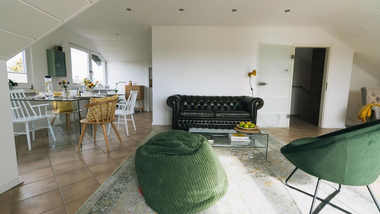 Een moderne woonkamer met een elegant zwart bank en een ronde fauteuil. De ruimte is licht en vriendelijk, met een eettafel en kleurrijke stoelen op de achtergrond.