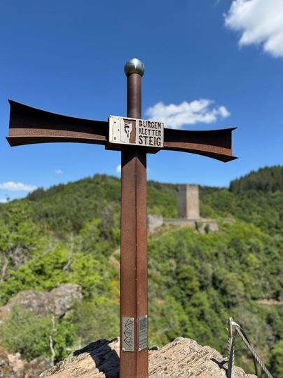 Een wegwijzer op een heuvel met uitzicht op een groene landschap en een kasteelruïne op de achtergrond. De lucht is blauw met enkele wolken.
