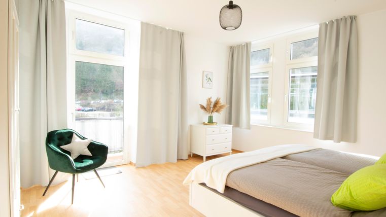 Ein helles Schlafzimmer mit großen Fenstern und einem bequemen Bett. Dazu eine gemütliche grüne Sitzecke und dekorative Pflanzen.