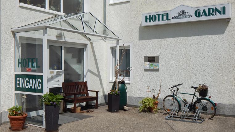 An inviting hotel entrance with a glass canopy. Next to the entrance door, there are plants and a bicycle.