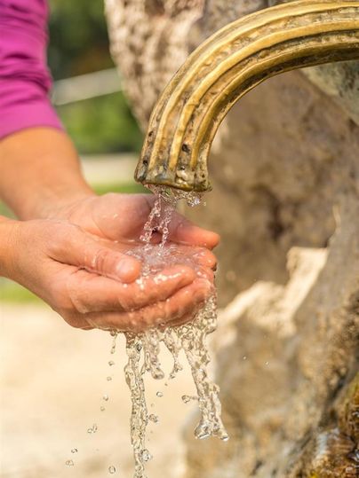 Hände fangen frisches Wasser aus einem antiken, bronzefarbenen Brunnen. Das Wasser sprudelt klar und lebendig.