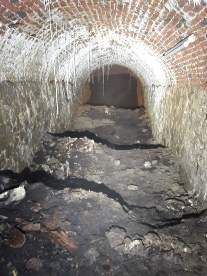 A dark tunnel with red brick walls and an uneven floor. A few slats and some dirt are scattered on the floor.