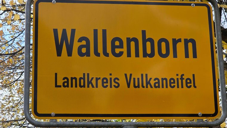 A yellow village sign with the inscription "Wallenborn" and "District Vulkaneifel". Surrounded by autumn leaves.