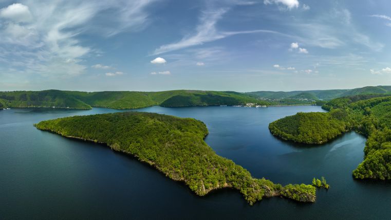 A picturesque lake surrounded by green hills and trees. The sky is clear with a few clouds.