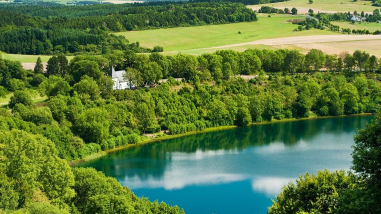 Ein malerischer See umgeben von üppigem Grün und sanften Hügeln. Der klare Himmel spiegelt sich im ruhigen Wasser.