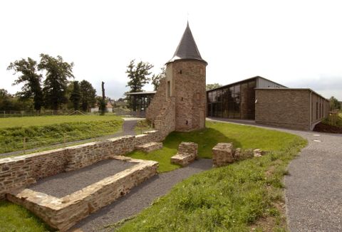 Een historische toren van steen met een spiraalvormig dak staat naast moderne gebouwen. De omgeving is groen met weilanden en bomen.