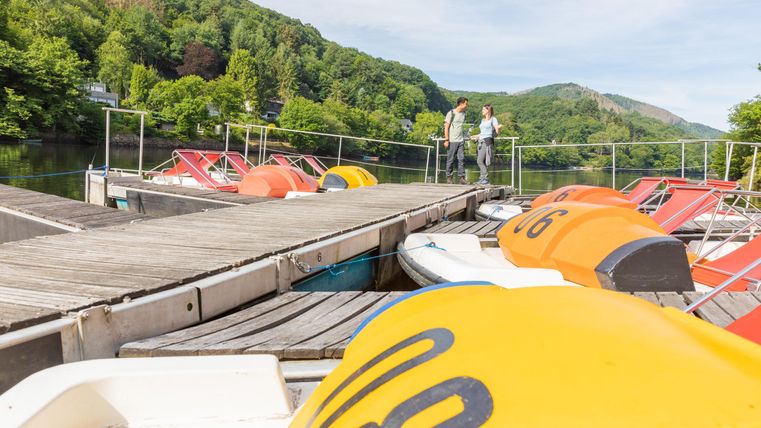 Eine malerische Boots-Anlegestelle mit bunten Tretbooten. Im Hintergrund sind grüne Hügel und zwei Personen, die am Ufer entlang gehen.