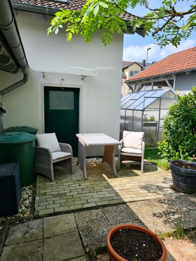 Ein gemütlicher Außenbereich mit einem Tisch und zwei Stühlen. Im Hintergrund sind ein Gewächshaus und grüne Pflanzen zu sehen.