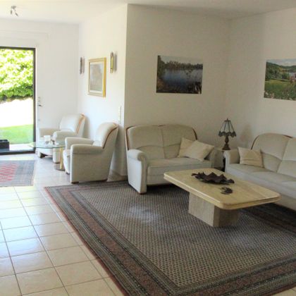A bright living room with white sofas and a table in the middle. Large windows offer a view of the garden.