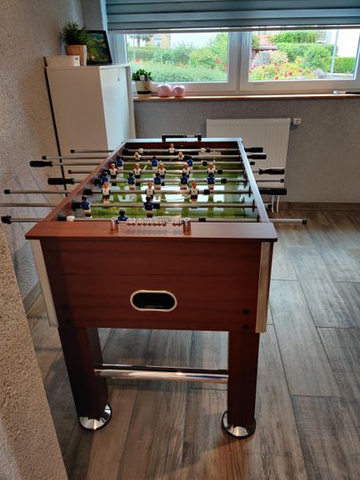 A modern foosball table made of wood stands in a room with a window. The surroundings are bright and friendly, ideal for games and leisure activities.