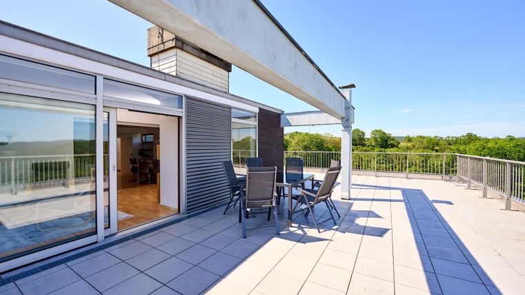 Eine moderne Dachterrasse mit einem Tisch und Stühlen. Im Hintergrund sind grüne Landschaften und ein klarer blauer Himmel zu sehen.