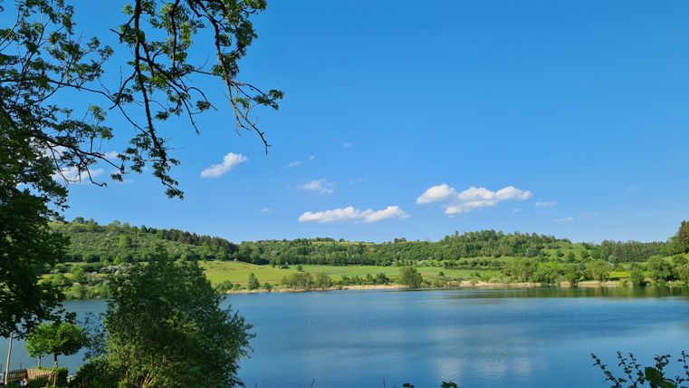 Ein ruhiger See umgeben von grünem Hügeln und Bäumen. Der Himmel ist klar und blau mit vereinzelten Wolken.