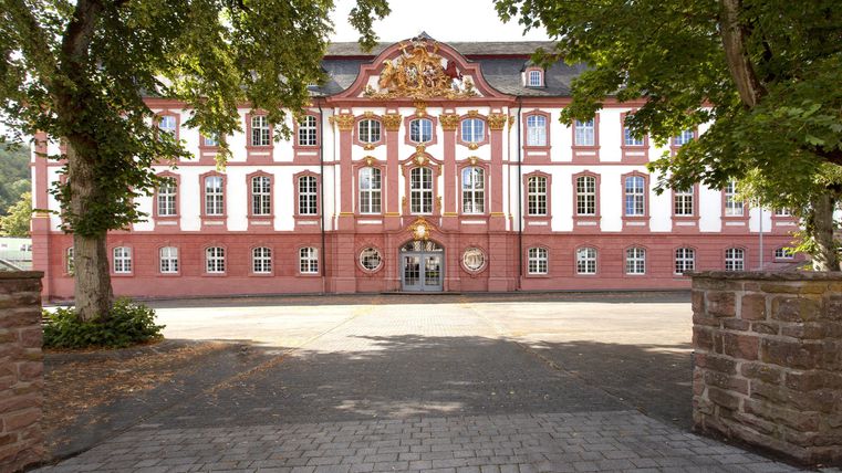 Ein großes, historisches Gebäude mit roter Fassade und weißen Fenstern. Bäume umrahmen den Eingangsbereich und verleihen der Szenerie eine grüne Note.