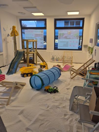 Een speelkamer met een zandvloer en verschillende speeltoestellen. Speelgoed zoals auto's en een tunnel zijn ook aanwezig.