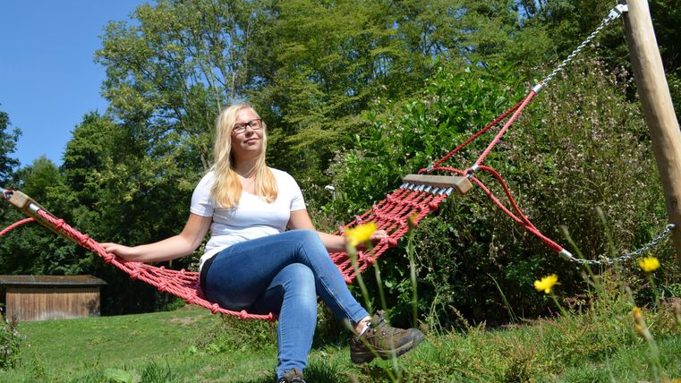 Eine Frau sitzt entspannt in einer roten Hängematte im Freien. Im Hintergrund sind grüne Bäume und eine blühende Wiese zu sehen.