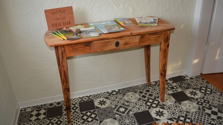 Ein Holzschirmtisch steht an der Wand, dekoriert mit Prospekten und einem Schild, das zum Eintragen ins Gästebuch einlädt. Der Boden ist mit einem bunten, gemusterten Fliesenbelag gestaltet.