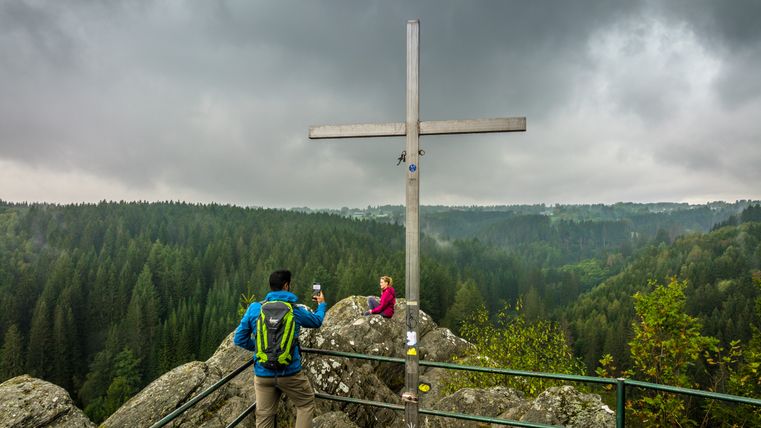 Two people at a viewpoint with a cross, surrounded by forest and a cloudy sky.