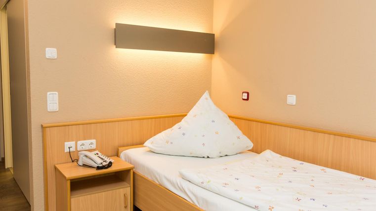 Een eenvoudige slaapkamer met een houten bed en een kussen in driehoeksvorm. De muren zijn in een neutrale kleur geschilderd, en de verlichting is zacht.