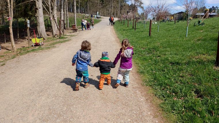 Drei Kinder halten Händchen und gehen auf einem Schotterweg. Um sie herum ist eine grüne Wiese und Bäume.
