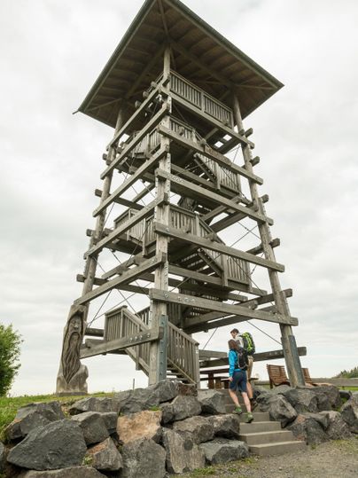 Ein hölzerner Aussichtsturm steht in der Natur. Zwei Wanderer nähern sich dem Turm auf einem steinigen Weg.
