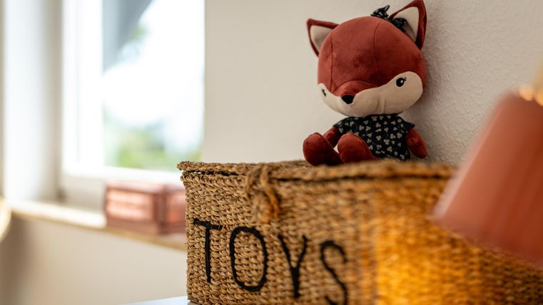 Ein niedlicher Plüschfuchs sitzt auf einem Korb mit der Aufschrift "TOYS". Der Korb steht in einem hellen Raum mit Fensterlicht.