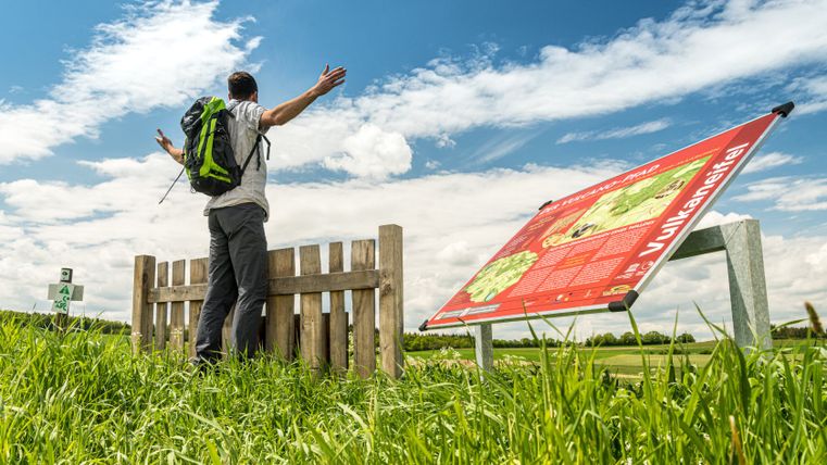 A hiker stands with outstretched arms in front of a wooden fence on the Vulcano Trail. An information sign is visible in the foreground.
