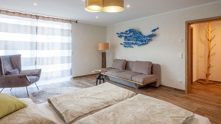 Ein modernes Wohnzimmer mit einer gemütlichen Couch und einem Sessel. Große Fenster sorgen für viel Tageslicht und eine entspannte Atmosphäre.