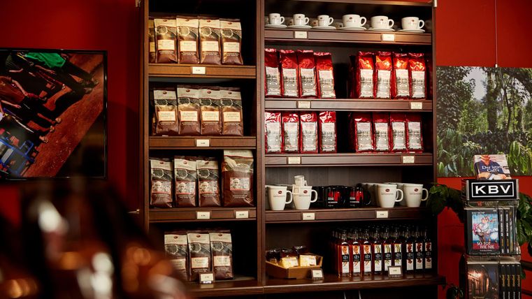 Ein gut sortiertes Regal in einem Café, voller Kaffeetüten und Tassen. Die Wände sind rot gestrichen, was eine einladende Atmosphäre schafft.