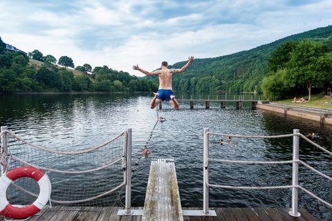 Ein Mann springt von einem Steg in einen See. Im Hintergrund sind grüne Hügel und ein bewölkter Himmel zu sehen.
