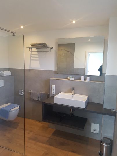 A modern bathroom with a shower, sink, and wooden floor. The wall design is neutral and well-kept.