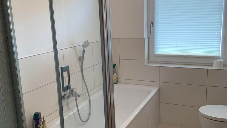 Een moderne badkamer met een douche en een bad. Er zijn grote tegels en een raam dat natuurlijk licht binnenlaat.