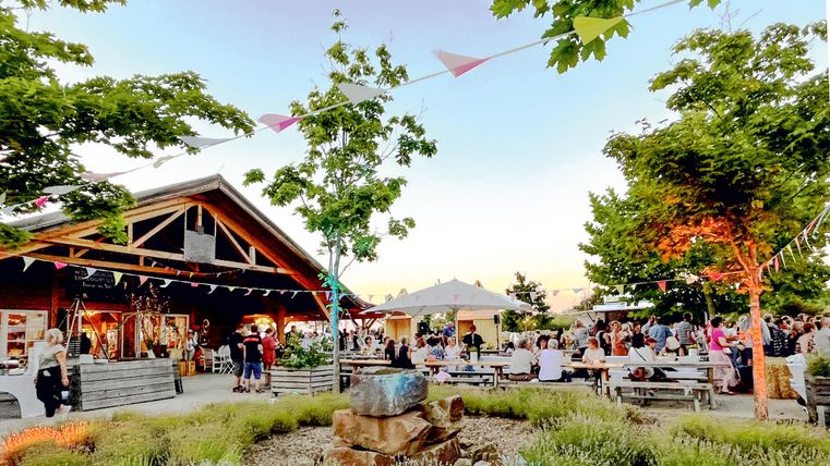 Een levendige buitenactiviteit met veel mensen die zich onder de bomen verzamelen. Op de achtergrond is een mooi gebouw te zien met feestelijke decoratie.