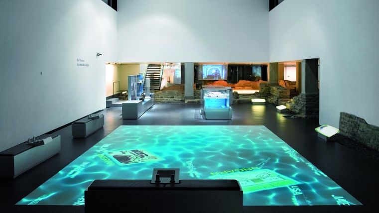 Ein modernes Ausstellungsraum mit Projektionen auf dem Boden, die Wasser darstellen. Auf der rechten Seite sind verschiedene Exponate und Vitrinen zu sehen.