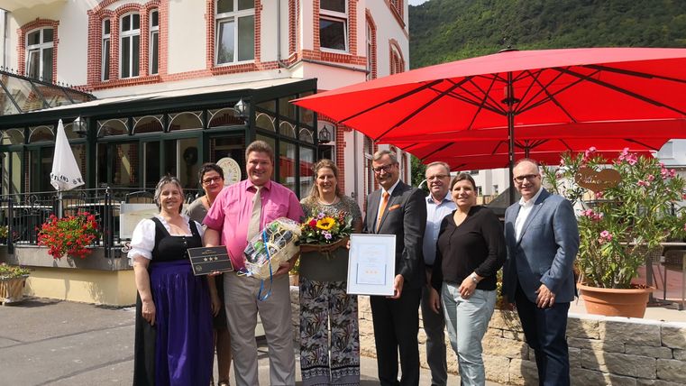 Eine Gruppe von Menschen steht vor einem Restaurant mit roten Sonnenschirmen. Sie lächeln und halten Auszeichnungen sowie Blumen in der Hand.