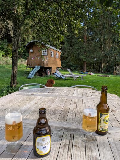 A table outside with three bottles of beer and glasses. In the background, there is a wooden house and there are lounge chairs on the grass.