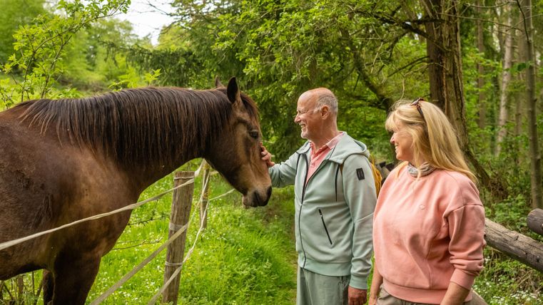 Een paar staat op een groene weg en aait een paard. Op de achtergrond zijn bomen en veel natuur te zien.