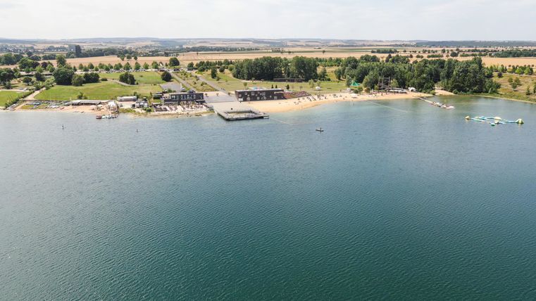 Ein ruhiger See mit einer Sandbucht im Vordergrund. Im Hintergrund sind Wiesen und Bäume zu sehen, ideal für entspannende Ausflüge.
