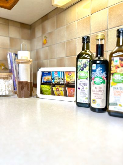 A modern kitchen with various bottles of olive oil on the countertop. In the background, numerous food items are visible in a chronicle box.