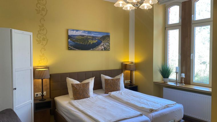 Ein gemütliches Schlafzimmer mit zwei Einzelbetten und warmen Farbtönen. Große Fenster lassen viel Licht herein, und es gibt dekorative Elemente wie ein Bild und Pflanzen.