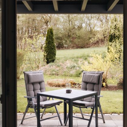 Ein gemütlicher Außenbereich mit einem Tisch und zwei Stühlen. Im Hintergrund ist ein schöner Garten mit frischem Grün zu sehen.