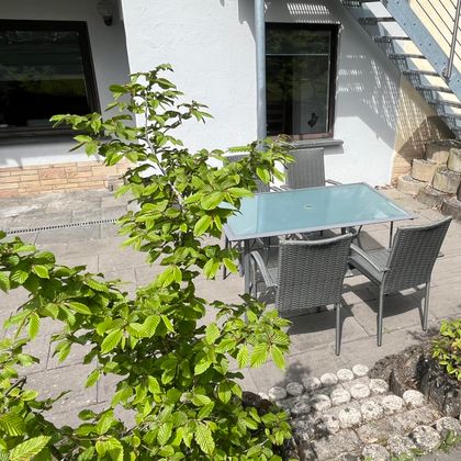 A cozy outdoor area with a glass table and four chairs. In the background, you can see a bright house and green foliage.