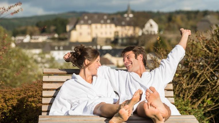 Ein Paar in Bademänteln entspannt auf einer Holzliege. Im Hintergrund sind sanfte Hügel und ein historisches Gebäude zu sehen.