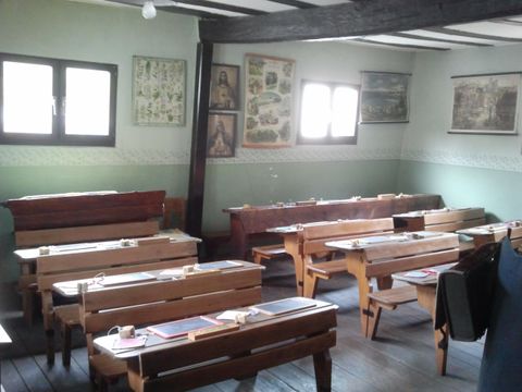 Ein Klassenzimmer mit Holztischen und Bänken. An den Wänden hängen historische Bilder und Karten.