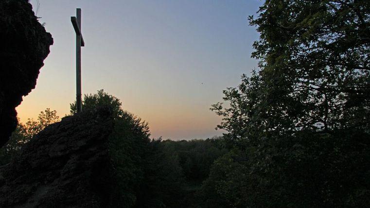 A wooden cross stands on a rock, surrounded by trees. The sky is painted in soft tones at sunset.